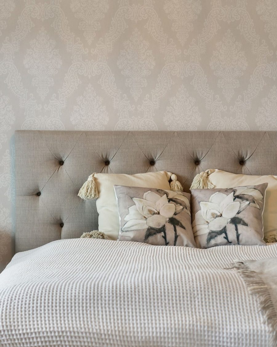 Calm neutral bedroom with a tone-on-tone damask wallpaper behind a tufted headboard and floral cushions