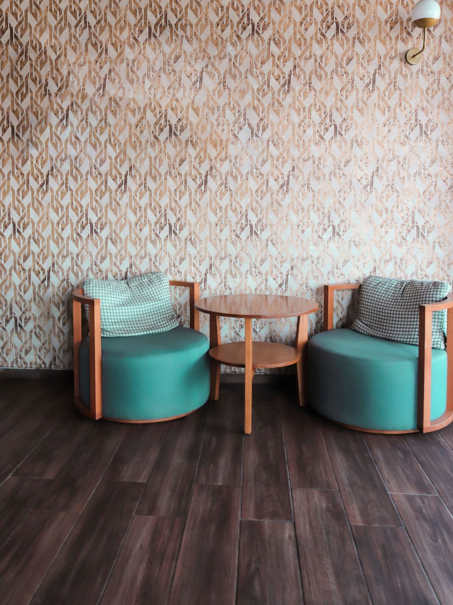 Sitting area with a warm terracotta and cream ogee-patterned wallpaper, two teal armchairs and a round wooden table