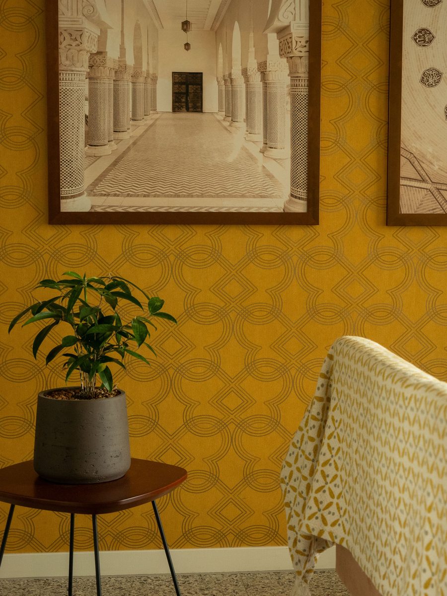 Mustard-yellow geometric wallpaper with a framed print, a potted plant on a side table and the edge of an armchair