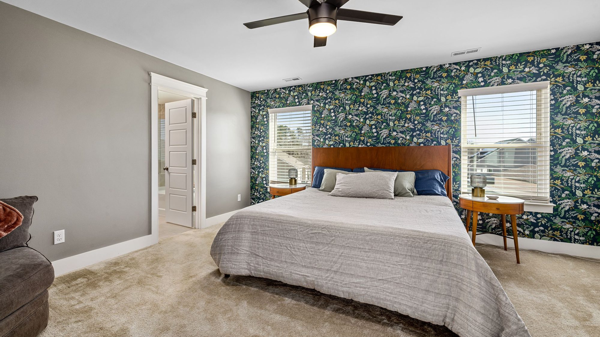 Bedroom with a deep green botanical wallpaper accent wall behind a wood-framed bed, two windows and a ceiling fan
