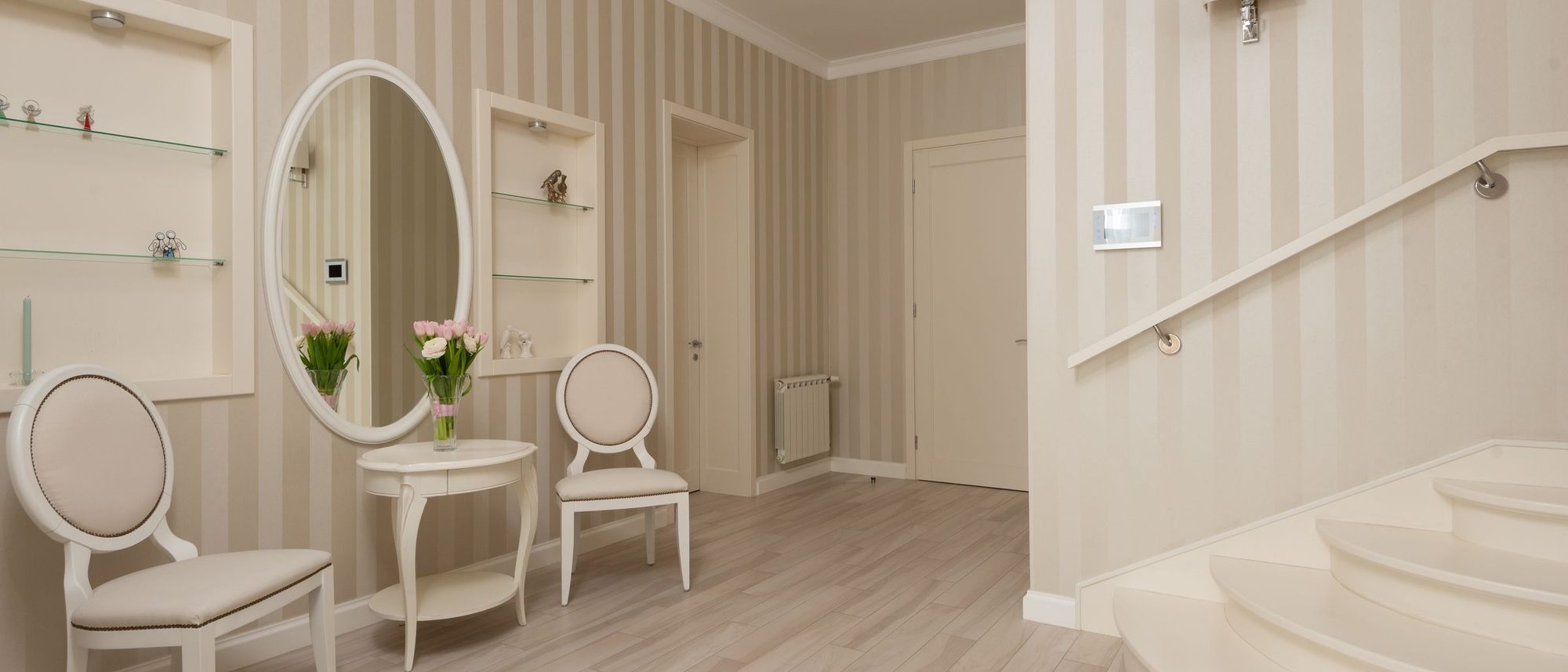 Light entry hall with a fine beige striped wallpaper, an oval mirror, white chairs and a staircase