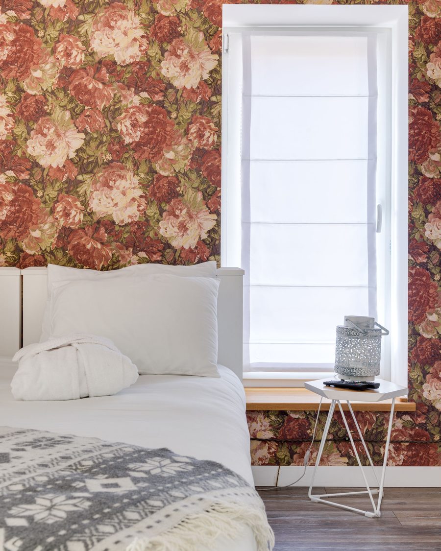 Bright bedroom with a bold red rose wallpaper around a window, white bedding and a small wire side table