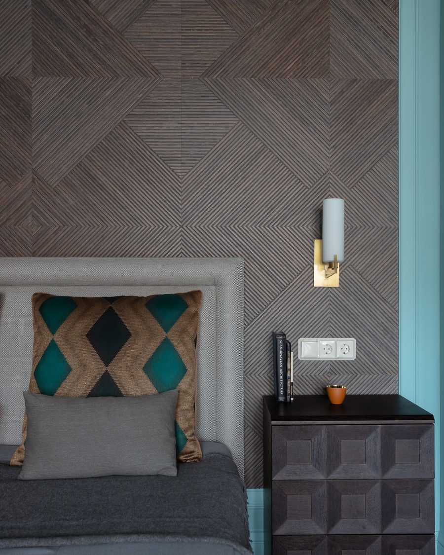 Textured wood-grain geometric wallpaper behind a grey upholstered headboard with a brass wall sconce